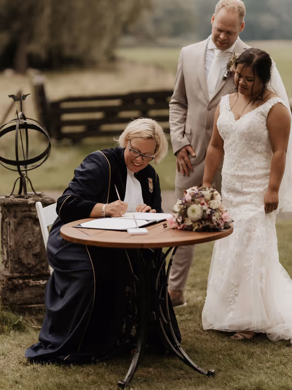 Trouwambtenaar zit achter tekentafel de akte te tekenen in groene toga en het bruispaar staat er naast